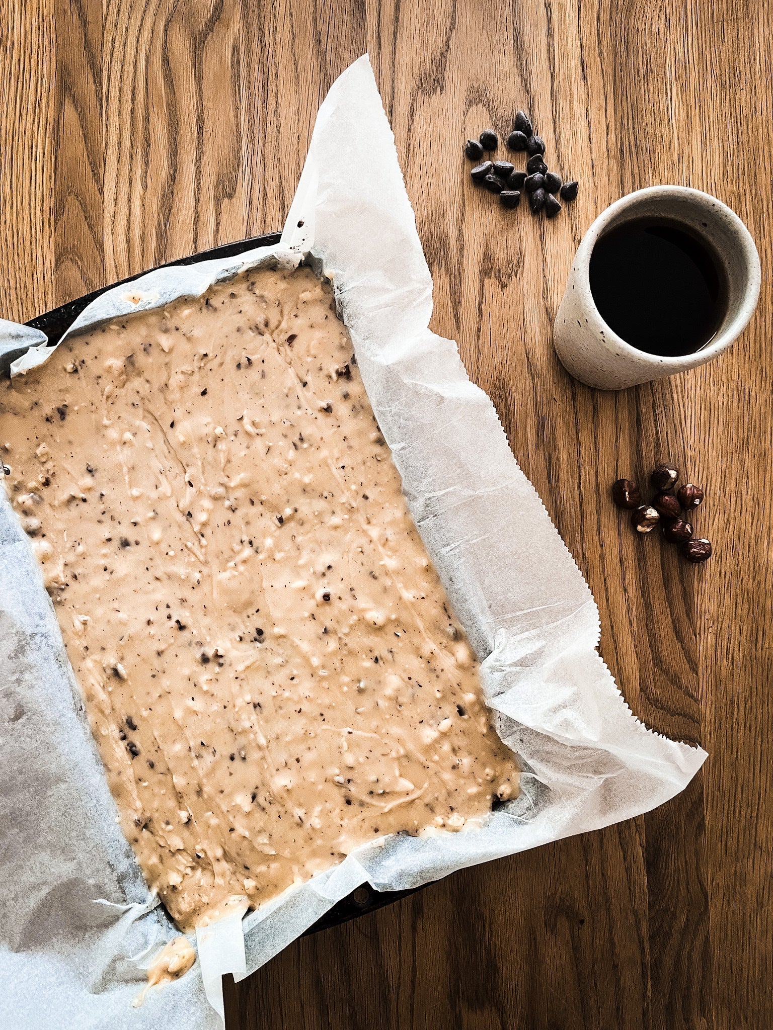A baking tin of Coffee Flapjacks on baking parchment on a wooden counter top beside a cup of coffee with hazelnuts and chocolate chips
