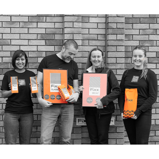 A black and white group shot of three women and one man against a brick wall background. Each person is holding something relating to FiXX coffee which is shown in vibrant orange.