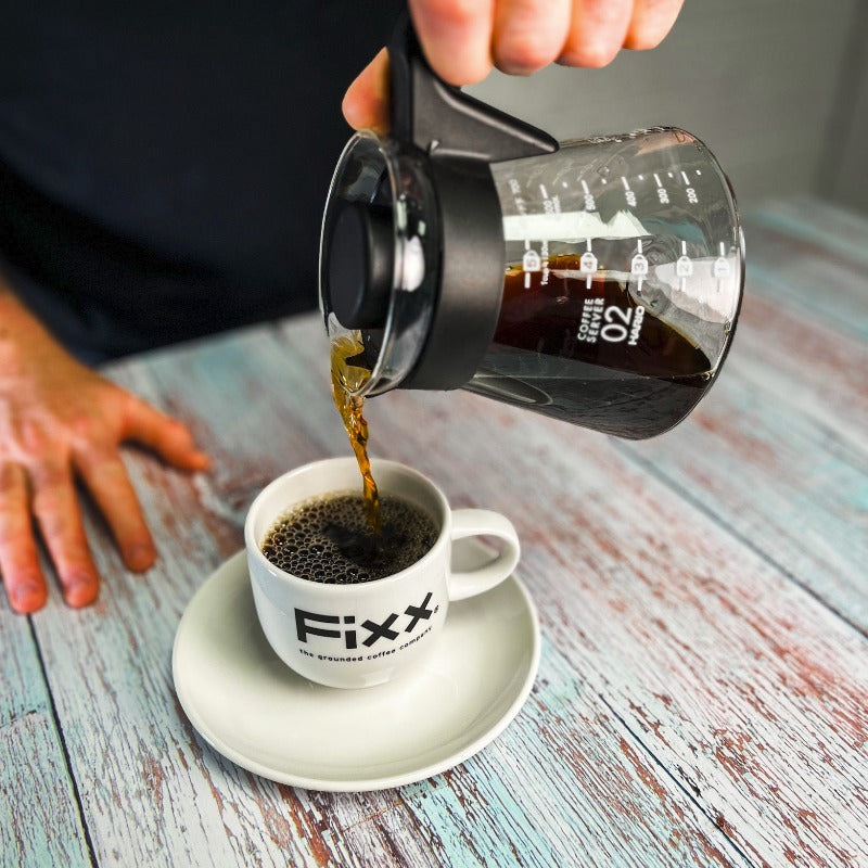 Person pouring coffee from a glass carafe into a white cup labeled &