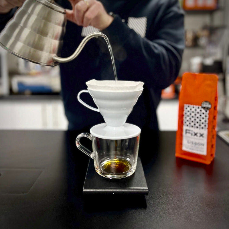 A person pouring coffee into a filter brewer on top of a glass cup on a black surface.