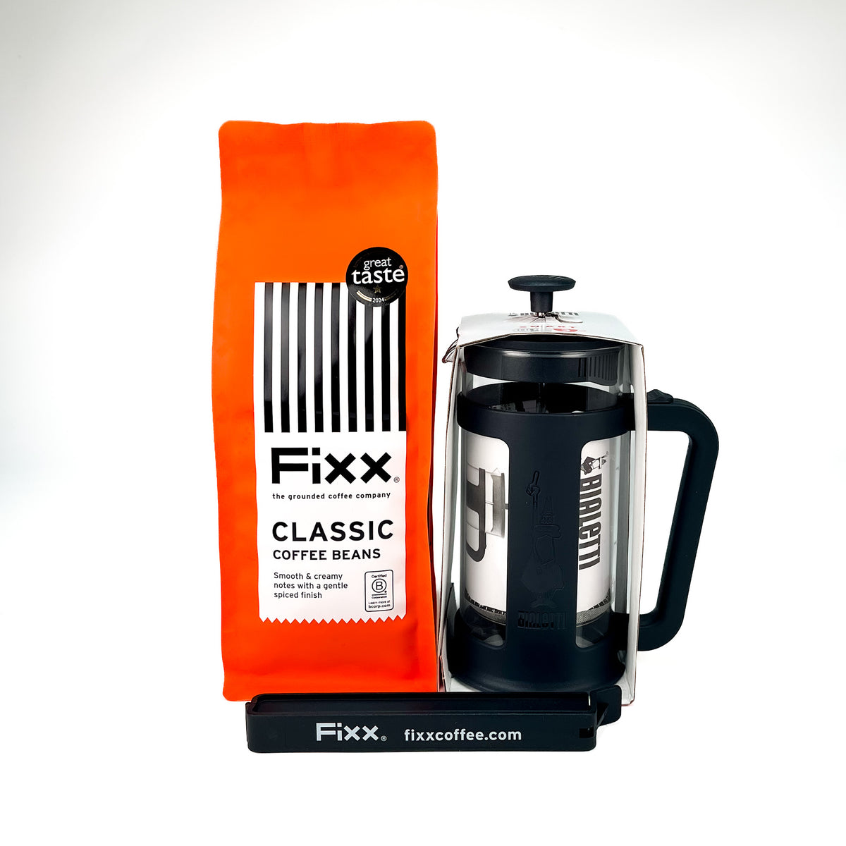 A bright orange bag of Fixx Classic Coffee Beans stands next to a black and stainless steel French press on a black branded base against a clean white background.