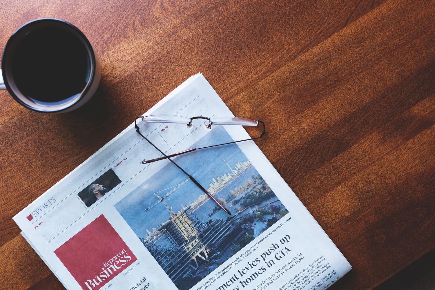 A wooden table top with a cup of black coffee, a newspaper and a pair of glasses.