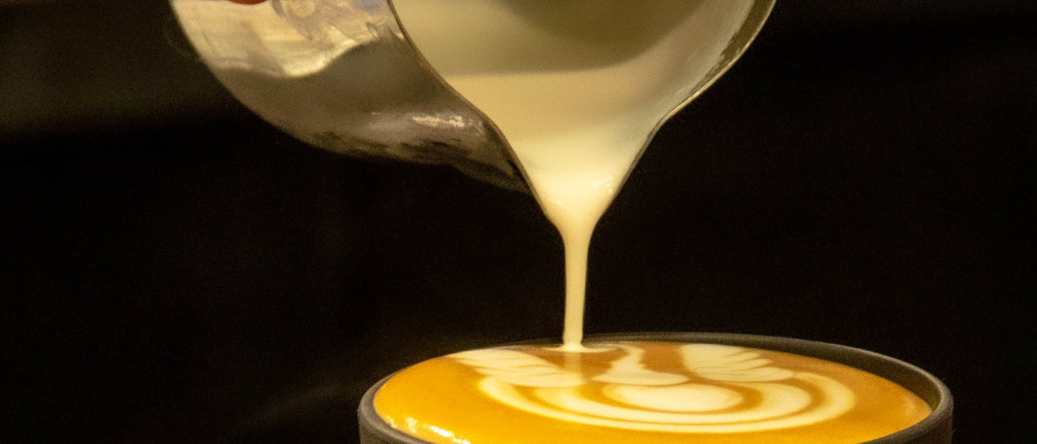 Milk slowly pouring from a metal jug making a latte art design in a cup of coffee