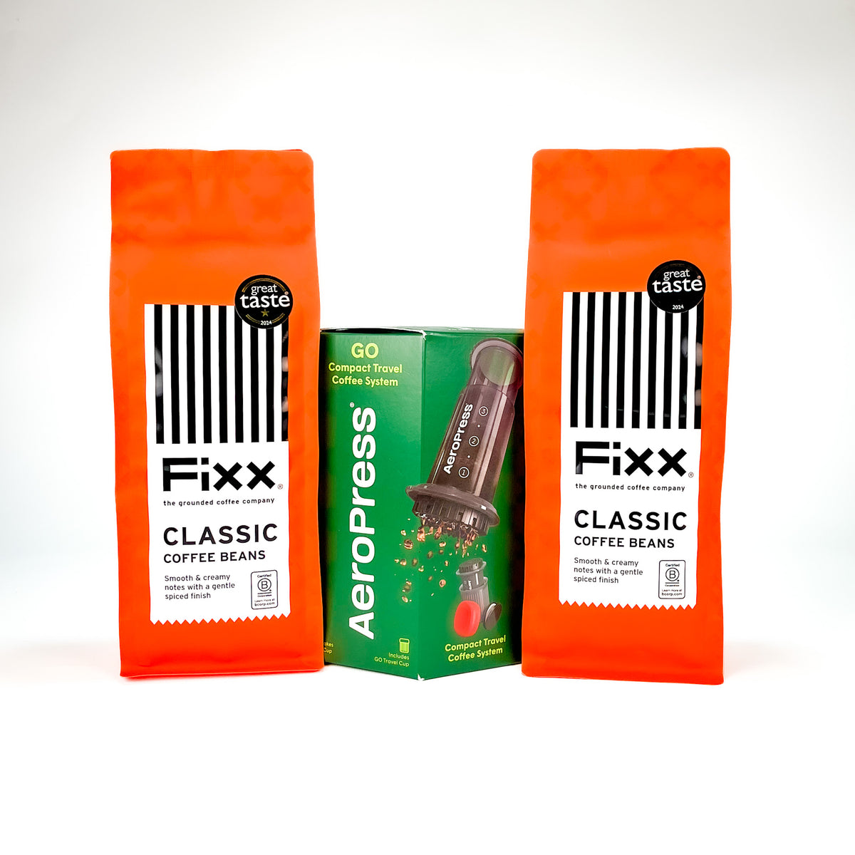 Two orange bags of Fixx Classic Coffee Beans stand on either side of a green Aeropress travel coffee system box, photographed on a clean white background.