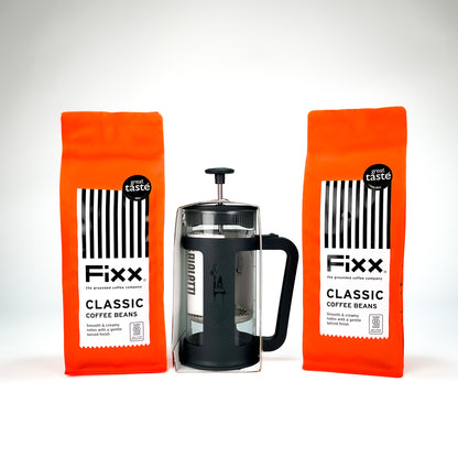 Two bright orange bags of Fixx Classic Coffee Beans flanking a black and glass French press, arranged symmetrically on a clean white background.