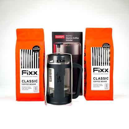 A coffee gift set featuring two bright orange bags of Fixx Classic Coffee Beans, a black Bouclier French press, and a black Boxburu electric coffee grinder, arranged on a white background.