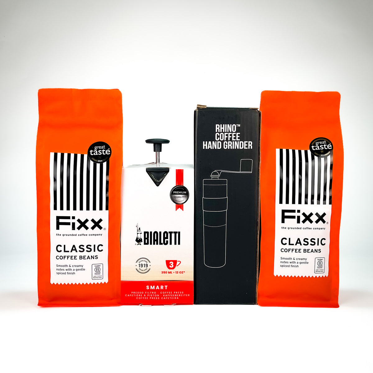 Three coffee-related products arranged against a light gray studio background: two bright orange Fixx coffee bean bags flanking a central white Bialetti coffee maker, with a black Rhino Coffee Hand Grinder box in the middle.