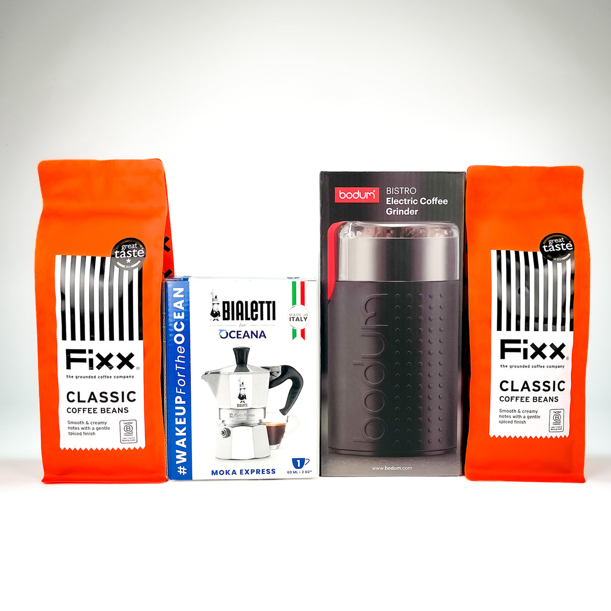 A product bundle featuring two orange bags of Fixx Classic Coffee Beans, a white and blue box of Bialetti Oceana Moka Express, and a black Bodum 31500 electric coffee grinder, arranged against a light gray background.