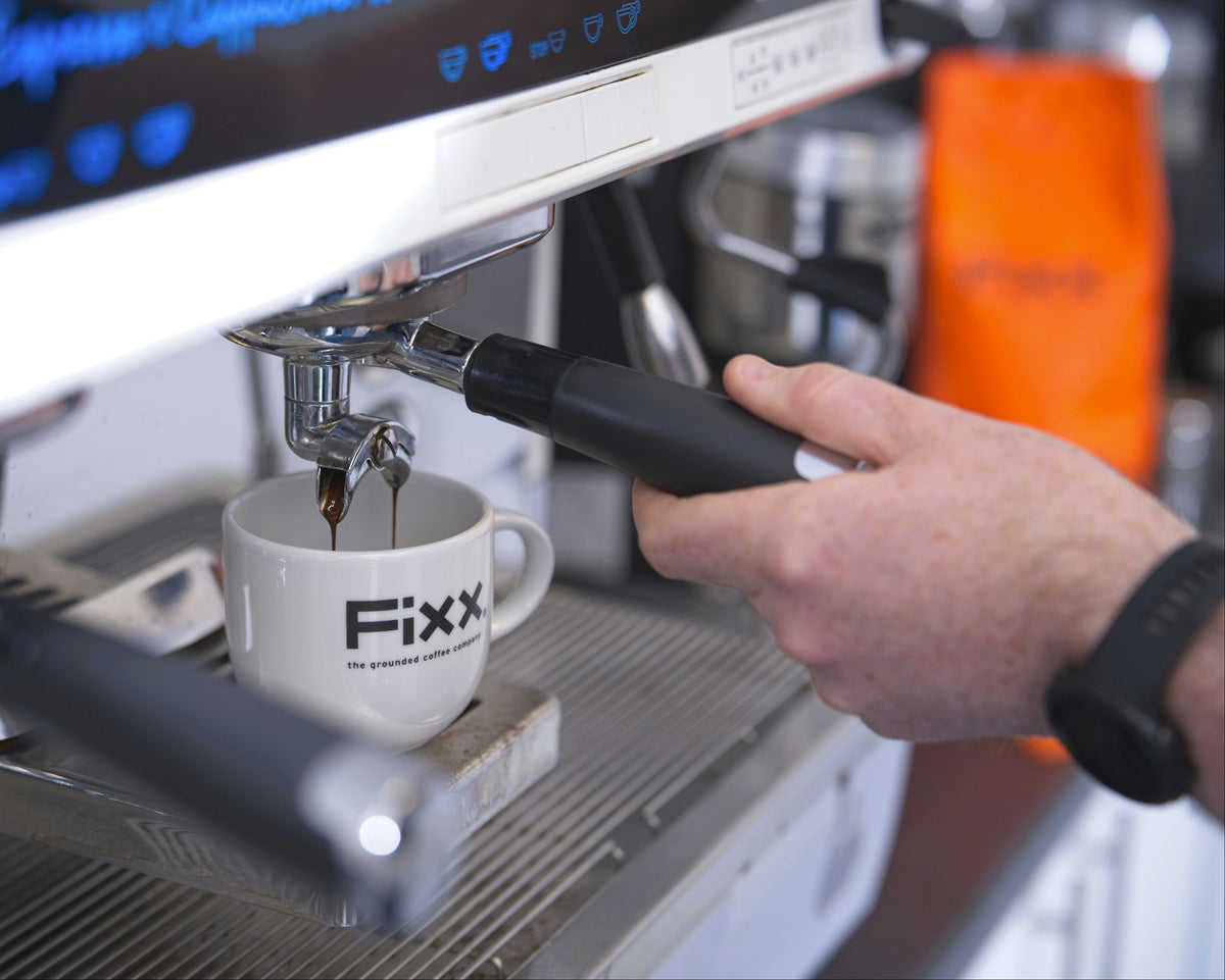 Espresso being brewed into a white cup with 'Fixx' branding, held by a hand above an espresso machine.