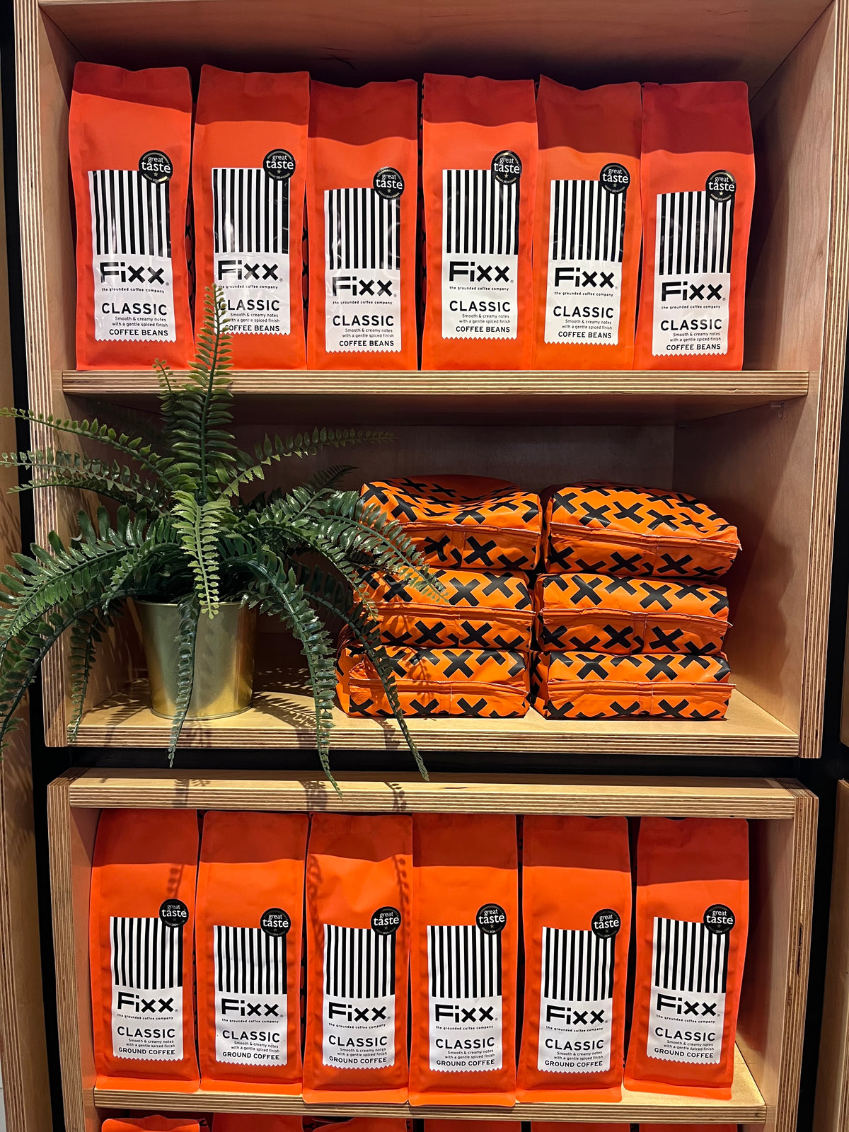 Multiple bags of Fixx Classic Coffee Roast arranged on wooden shelves, with one green potted fern and a stack of patterned orange-and-black fabric items on the middle shelf.