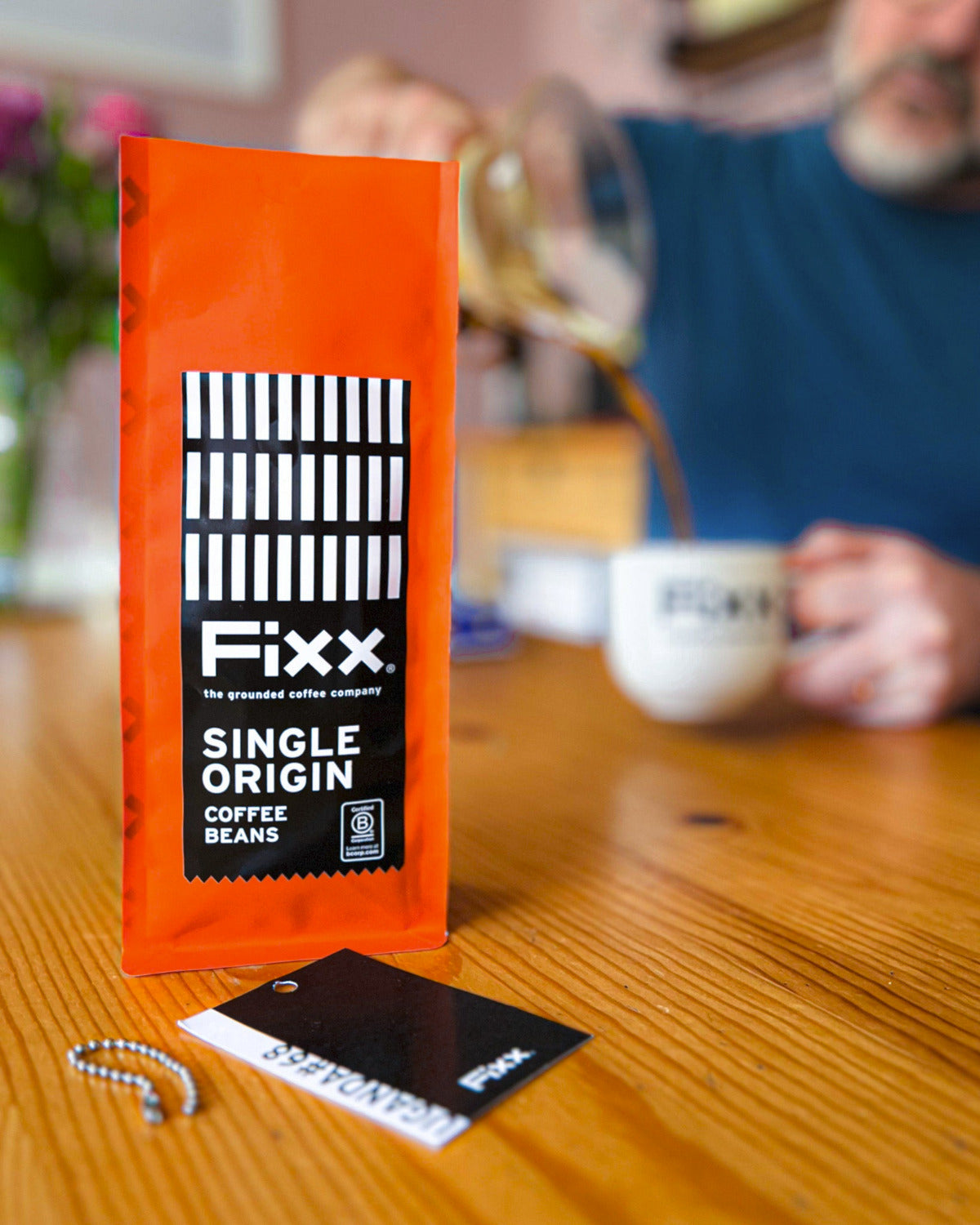 An orange bag of Fixx Single Origin Coffee Beans stands on a wooden table in a cafe setting, with a person in the background pouring coffee into a cup.