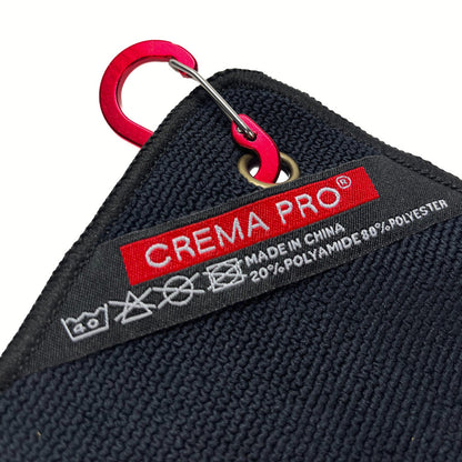 Close-up of a black textured fabric item with a red and black CREMA PRO label and a bright red carabiner attached, on a white background.
